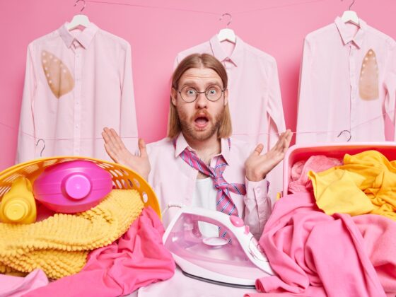 Overwhelmed bearded redhead man embarrassed as has much work to do irons laundry at home stands near